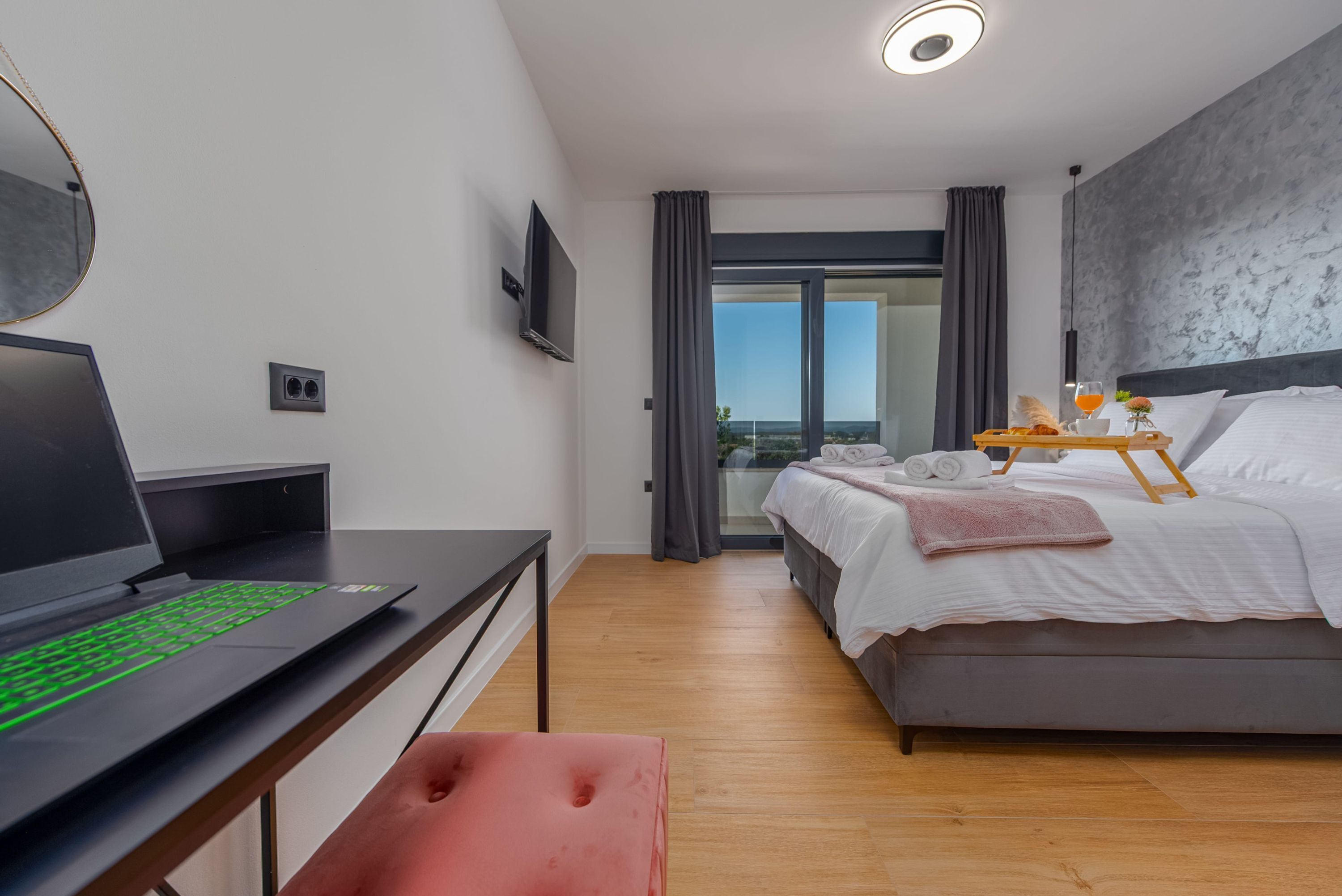 A modern hotel room with a bed, a desk with a laptop, a TV, and a balcony with a view.