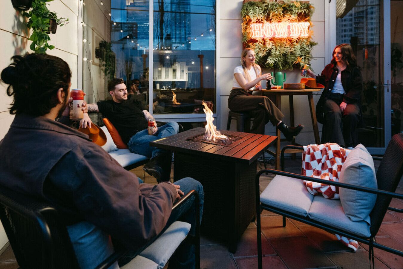 A group of people seated around a table with a fire, in an outdoor setting with a city view.
