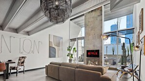 A modern living room with a large sectional sofa, a dining area with a table and chairs, a large window with a view of buildings, and a Netflix sign on the wall.