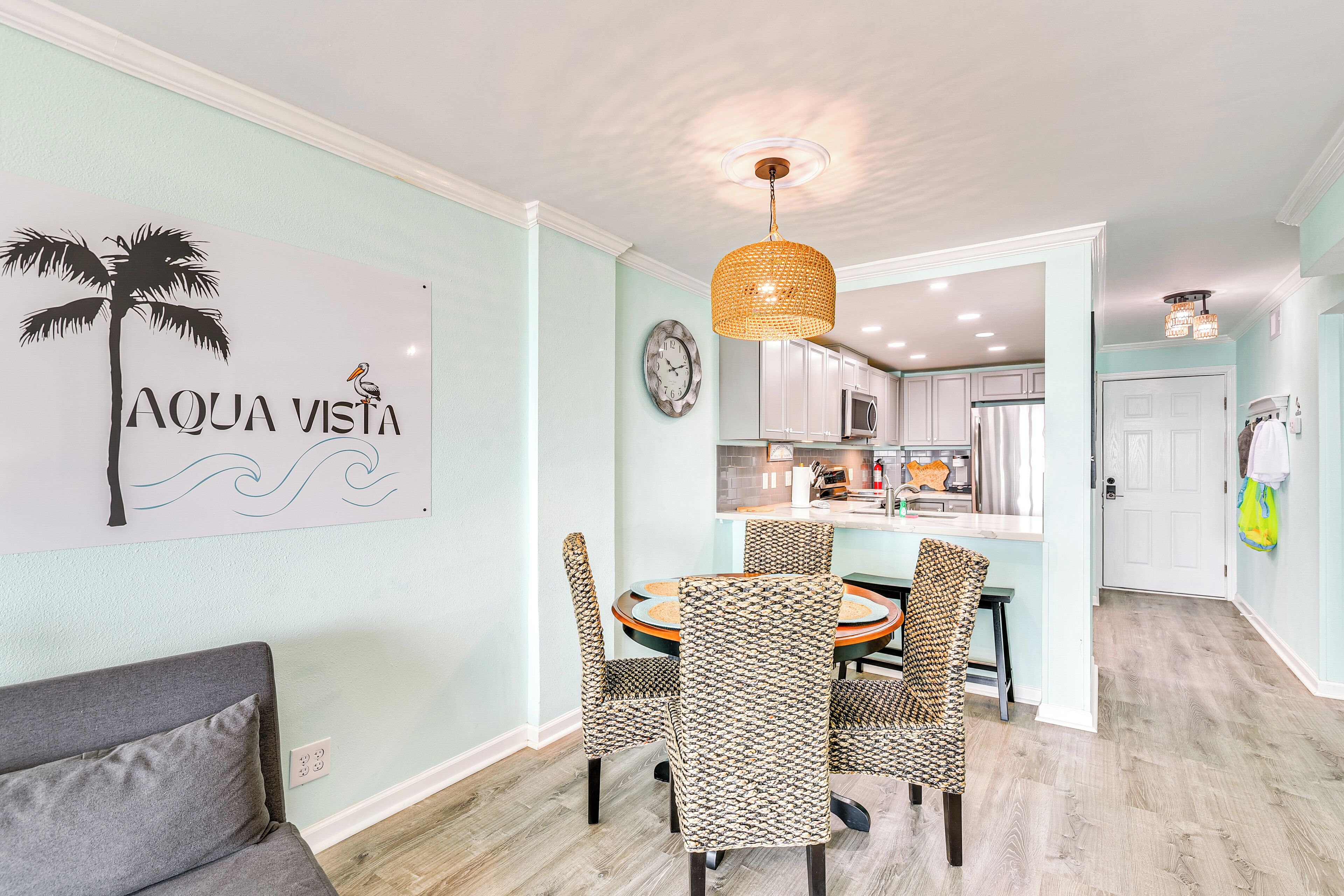 A dining area with a round table and patterned chairs, a wall clock, and a kitchen in the background.