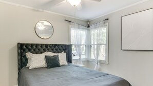 Ein Schlafzimmer mit einem gepolsterten Kopfteil, einem großen Bett mit grauer Bettwäsche, einem runden Spiegel und einem Fenster mit durchsichtigen Vorhängen.