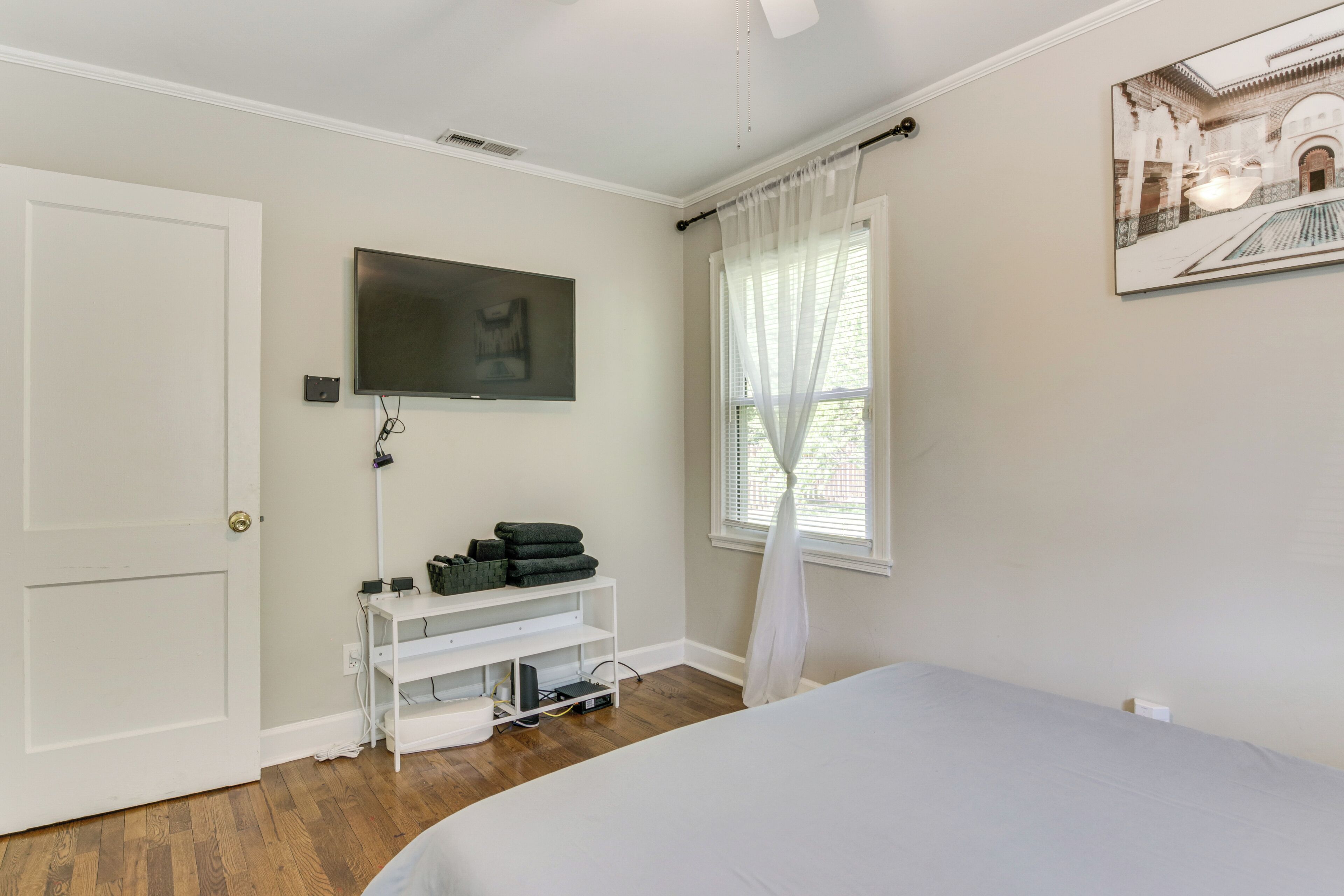 A bedroom with a bed, a TV mounted on the wall, a window with sheer curtains, and a white door.