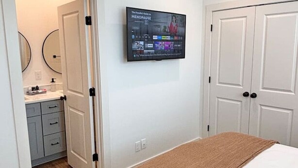 Un dormitorio con cama, televisor montado en la pared, armario y baño visible a través de una puerta abierta.