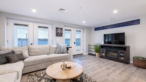 A modern living room with a large sofa, a coffee table, and a television. There are large windows with a view of a lake.
