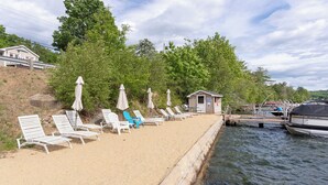 A waterfront property with a sandy beach, lounge chairs, umbrellas, and a small cabin.