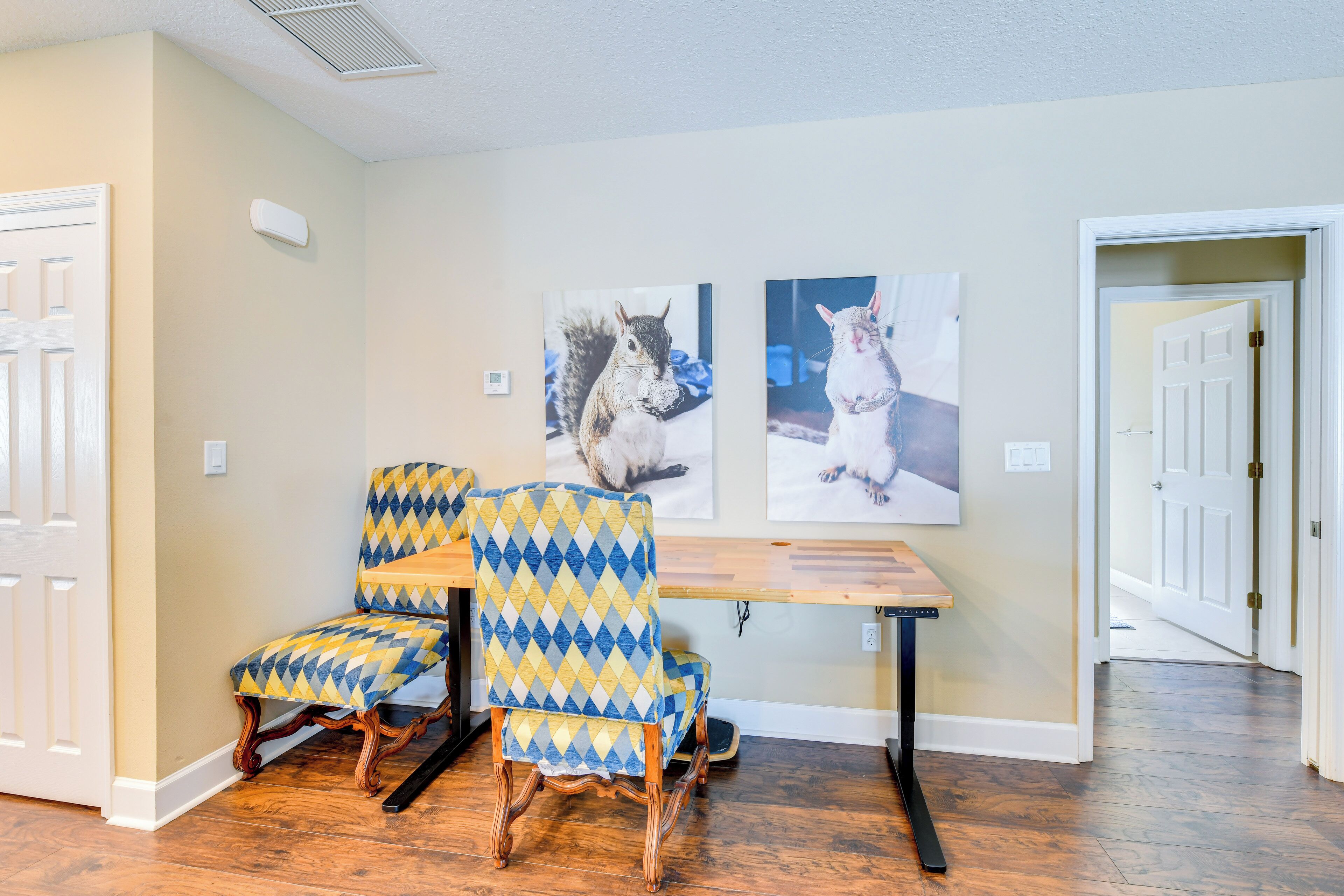 Une pièce avec un plancher en bois, une table à manger, deux chaises recouvertes d’un tissu à motifs et deux tableaux représentant des animaux, accrochés au mur.