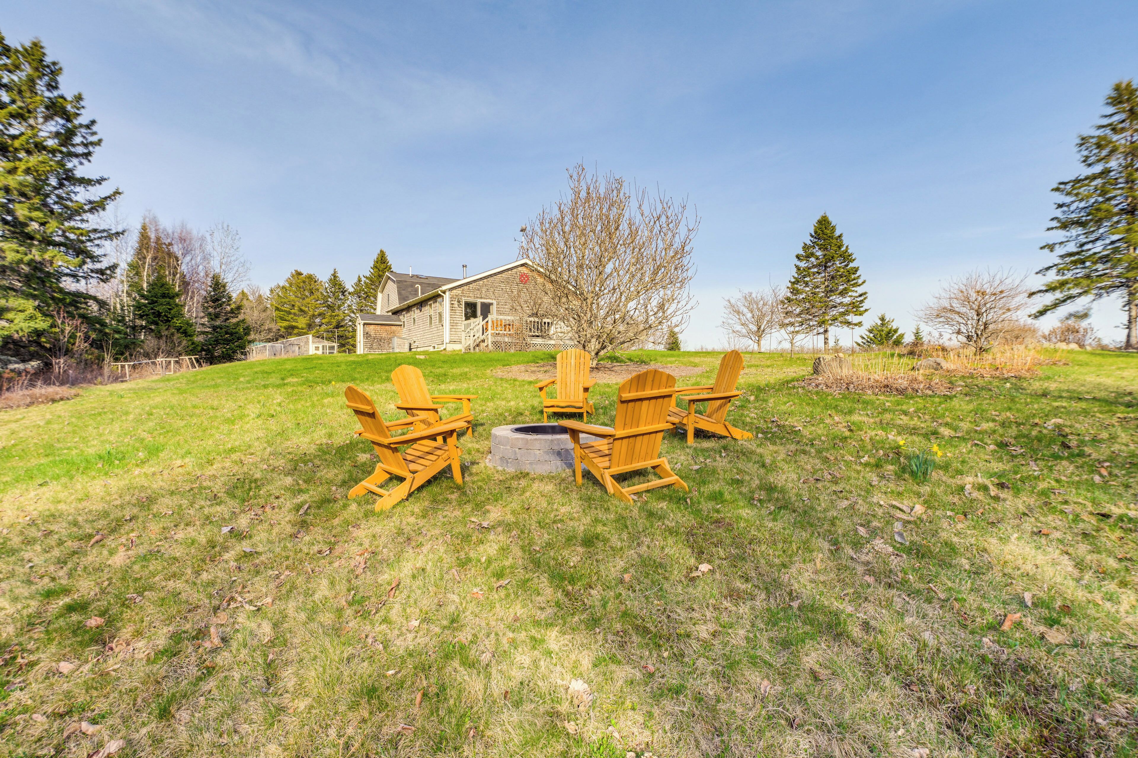 A grassy backyard with Adirondack chairs arranged around a fire pit, a stone house in the background, and a clear sky.