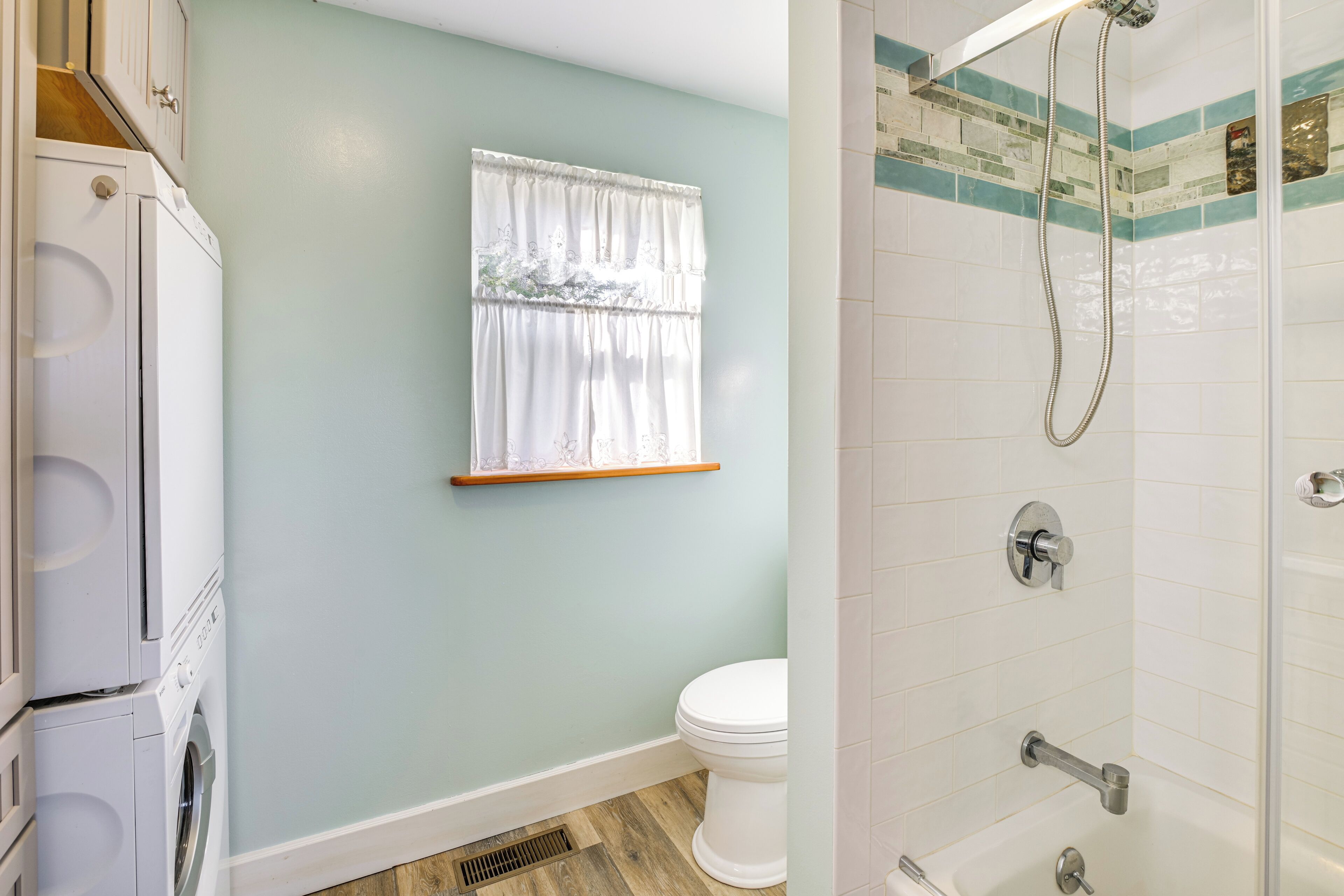 Un baño con un inodoro blanco, una ventana con cortina blanca, una ducha con cabezal de ducha manual y una pared con azulejos.