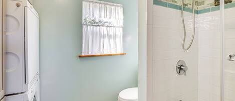 Un baño con un inodoro blanco, una ventana con cortina blanca, una ducha con cabezal de ducha manual y una pared con azulejos.