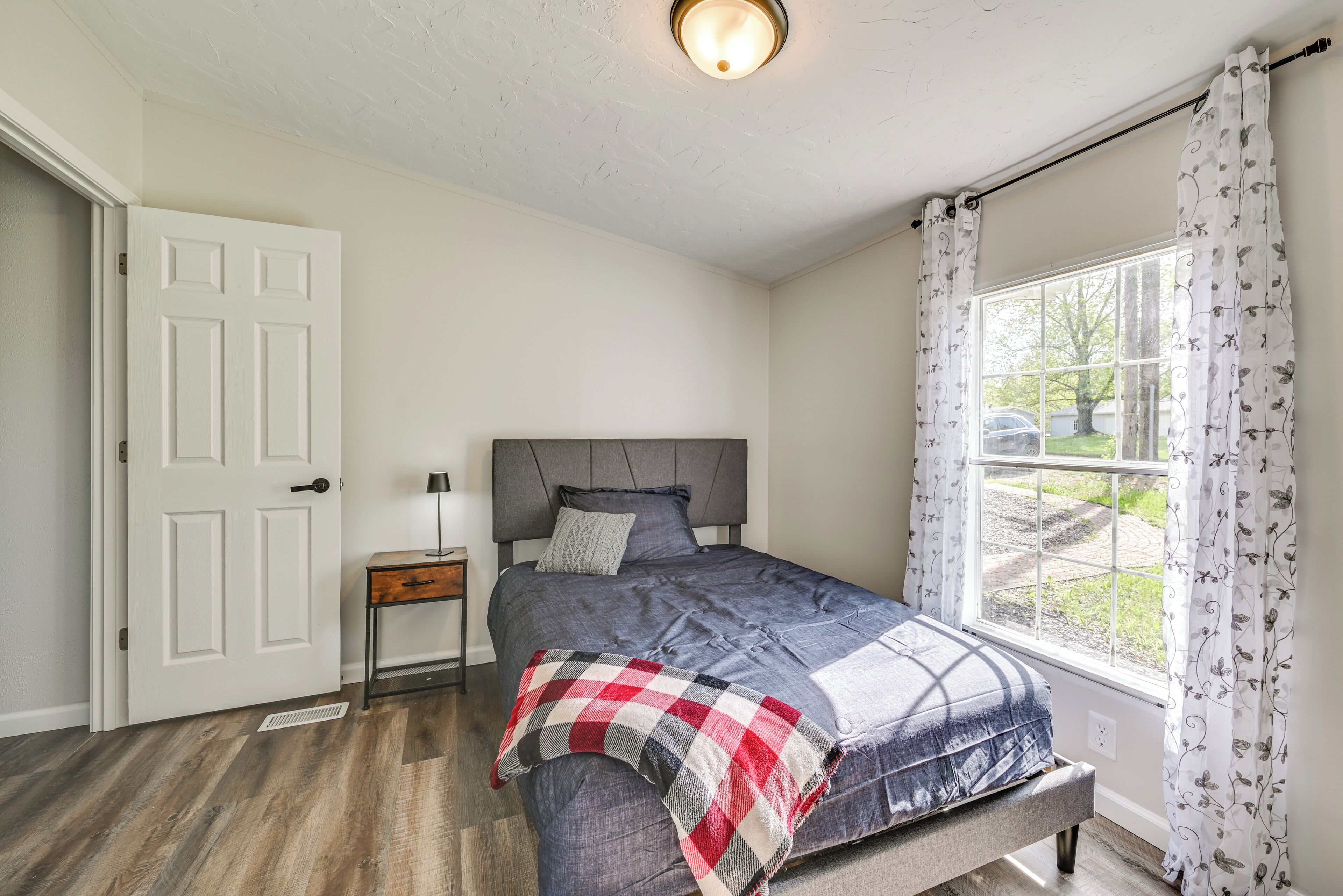 Un dormitorio con una cama, una mesita de noche, una ventana con cortinas y una puerta.