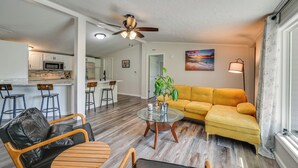 A modern living room with a yellow sofa, a glass coffee table, and a black leather chair. There is a kitchen area with white cabinets and a microwave. A ceiling fan is visible, and a painting hangs on the wall. - Near Hiking & Fishing: Dale Home w/ Deck! (Dale)