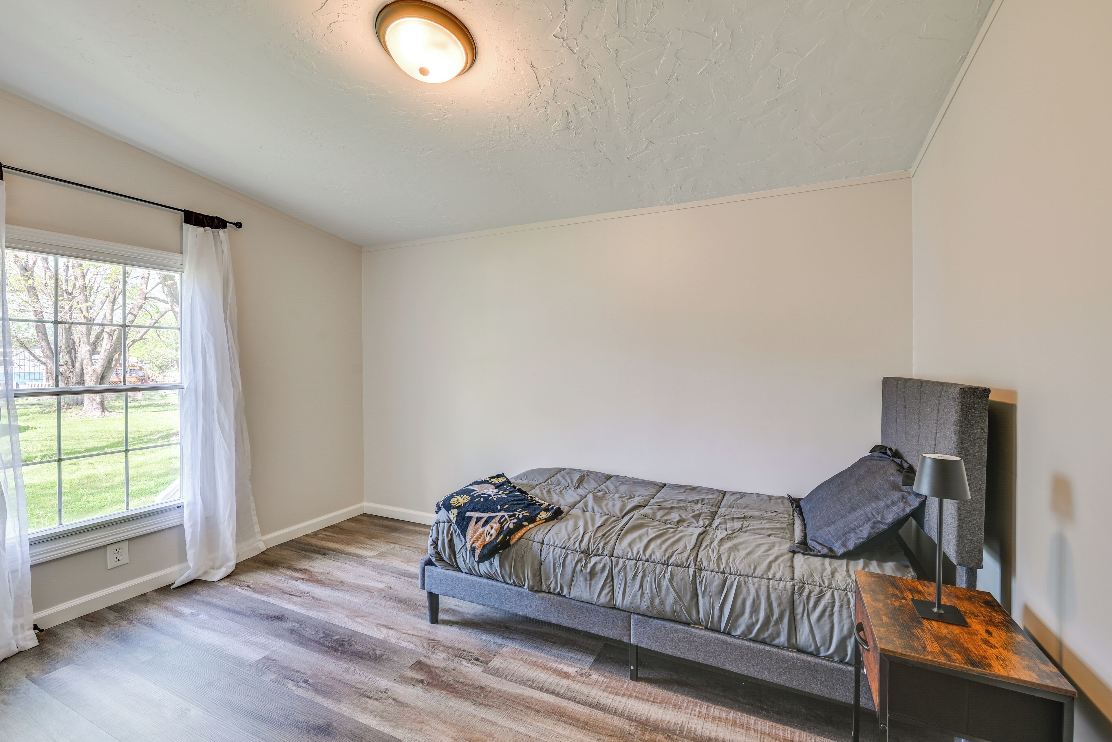 A bedroom with a bed, a nightstand, a lamp, a window with curtains, and a ceiling light.