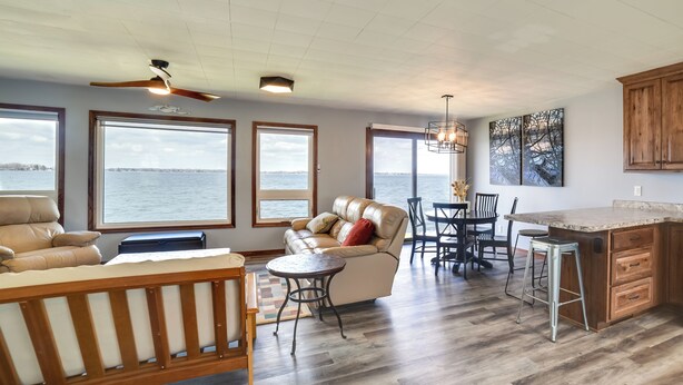 A spacious living area with a large window overlooking a body of water, a cozy seating area with a sofa and chairs, a wooden dining table, and a kitchen with wooden cabinets and a bar area. - BBQ Ready: Lakefront Retreat in Watertown (Watertown)