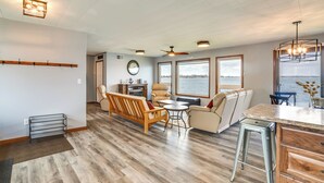 A spacious living area with a wooden floor, a sofa, a chair, a dining table, and a view of the water through large windows. - BBQ Ready: Lakefront Retreat in Watertown (Watertown)