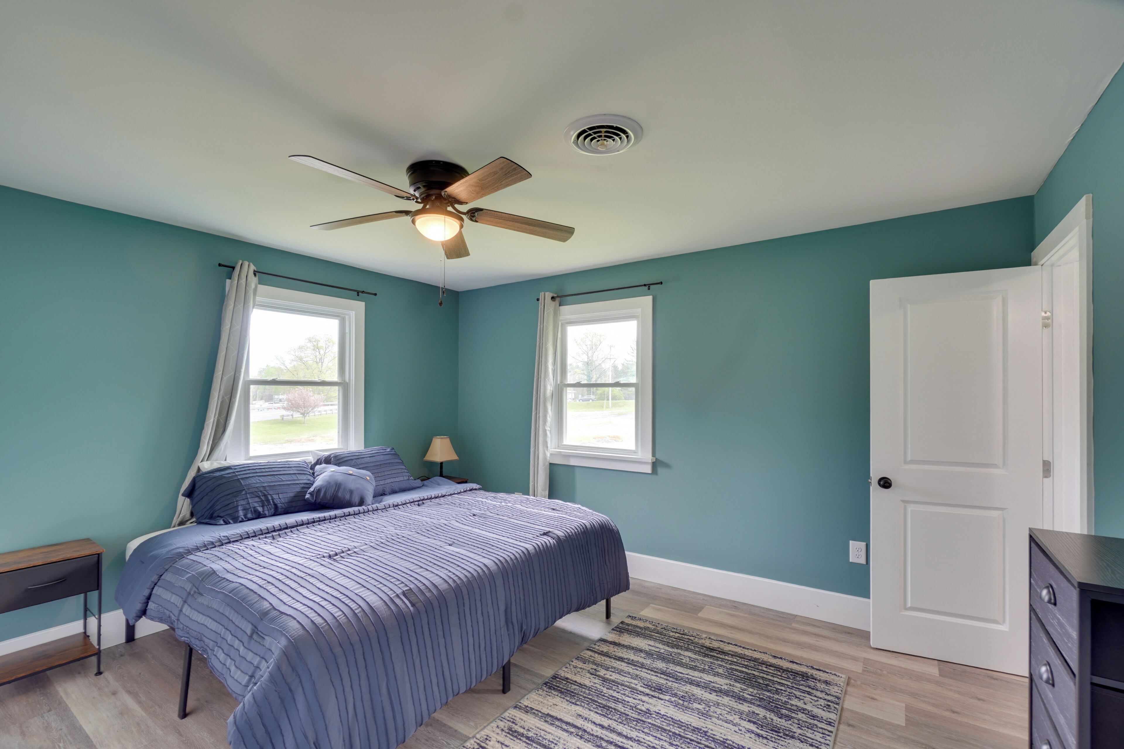 A bedroom with teal walls, a bed with a striped blue bedspread, a ceiling fan, two windows with white curtains, a wooden floor, and a white door.