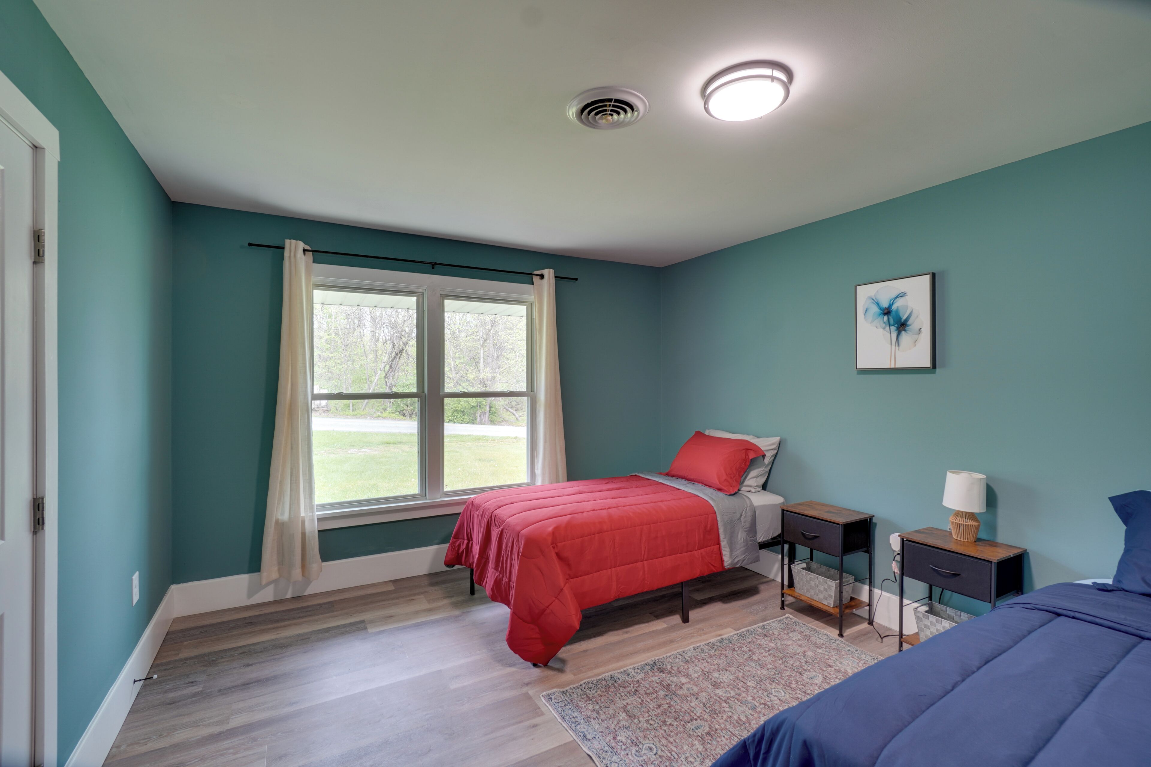 Un dormitorio con paredes color esmeralda, una cama con ropa de cama roja, una mesita de noche con lámpara y una ventana con cortinas.