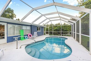 A covered swimming pool with a glass ceiling, surrounded by a blue house exterior, and equipped with a ladder and lounge chairs.