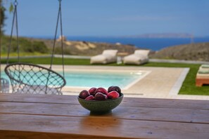 A wooden table with a bowl of fruit in the foreground, a pool, and lounge chairs in the background. - Oliva Emerald Eco - Secluded Off-grid Vineyard Villa (Hersonissos)