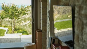 A room with a view of a grassy area and a tree, a stone wall, and a window with a view of a paved area. - Oliva Emerald Eco - Secluded Off-grid Vineyard Villa (Hersonissos)