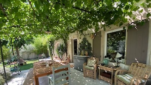 An outdoor patio area with a wooden table and wicker chairs, surrounded by greenery and a stone wall. - Charming Villa With AC in Alpilles (Paradou)