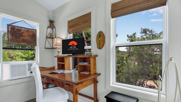 Une pièce confortable avec un bureau en bois, une télévision et une grande fenêtre donnant sur des arbres et un véhicule rouge à l’extérieur.