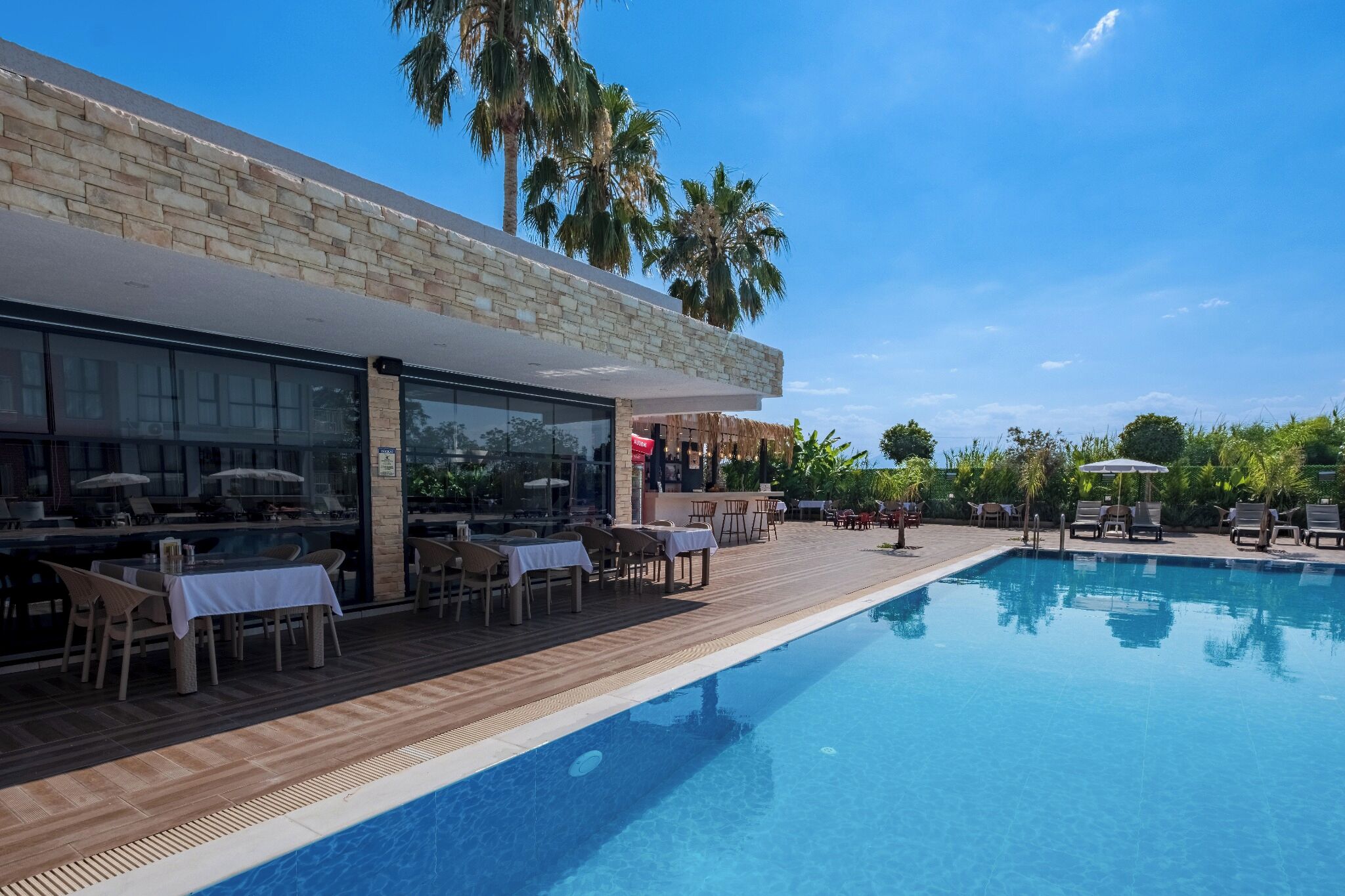 Zona junto a la piscina con mesas y sillas, piscina y un edificio de fachada de piedra y ventanas amplias.