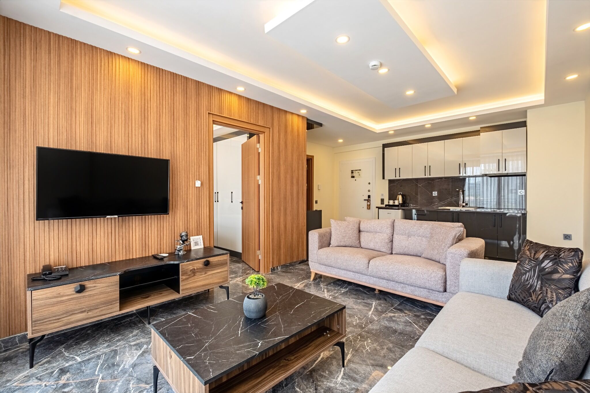Una sala moderna con un televisor de pantalla plana, un mueble de entretenimiento de madera, una mesa de centro de mármol, un sofá gris y una zona de cocina con armarios y un refrigerador.