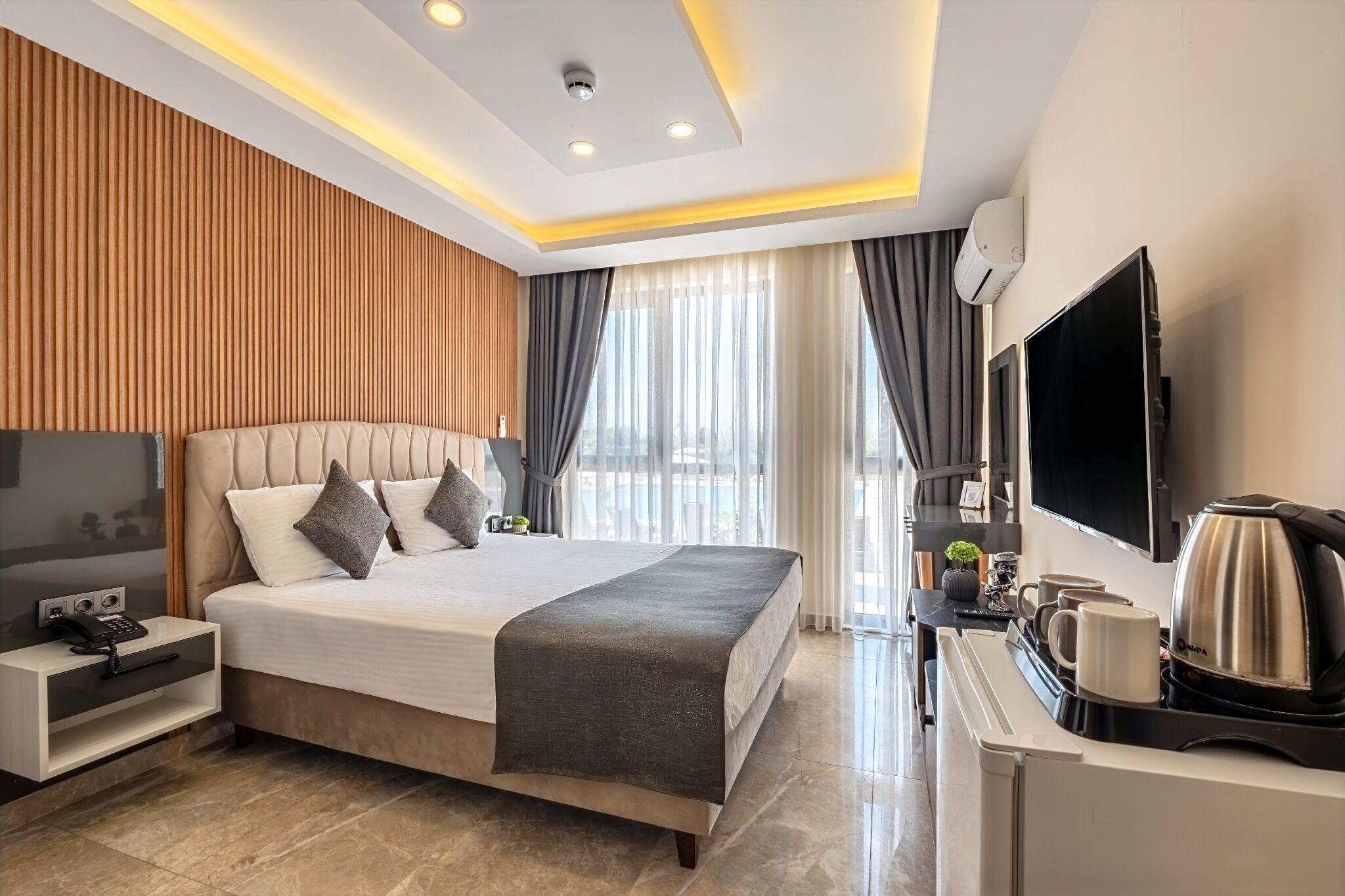 A modern hotel room with a large bed, bedside table with a phone, a flat-screen TV, a coffee maker, and a view of the outdoors.