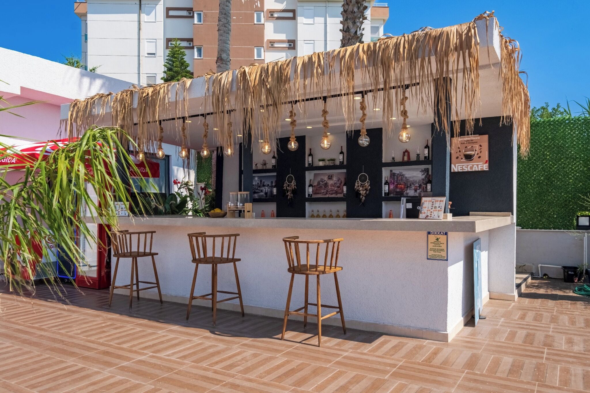 Un área de bar al aire libre con techo de paja, tres taburetes de madera y una barra con una pizarra de menú.
