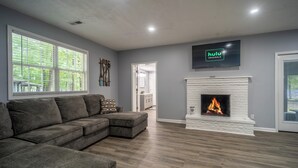 Una sala de estar con un sofá seccional gris, una chimenea blanca y un amplio ventanal con persianas.