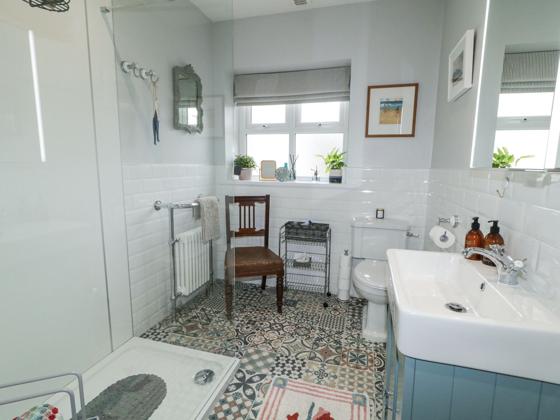 A bathroom with a shower, a toilet, a sink, and a chair. There are plants on the windowsill and a mirror on the wall.