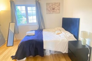 Une chambre à coucher avec un lit, une fenêtre avec des rideaux, une œuvre d’art accrochée au mur et une table de chevet avec une lampe.