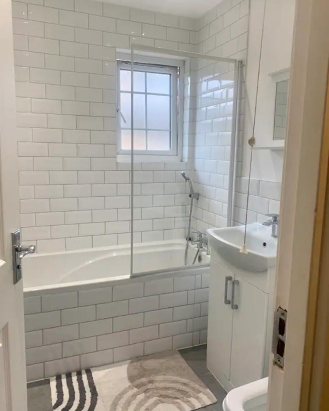 Een badkamer met witte metro betegeling, een bad, een douche, een wastafel en een raam.
