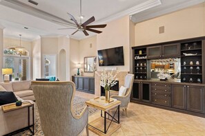 A living room with a sofa, armchairs, a coffee table, a TV, and a bar area with shelves and glasses. - Mediterranean Home with Club Amenities & Golf Cart (Destin)