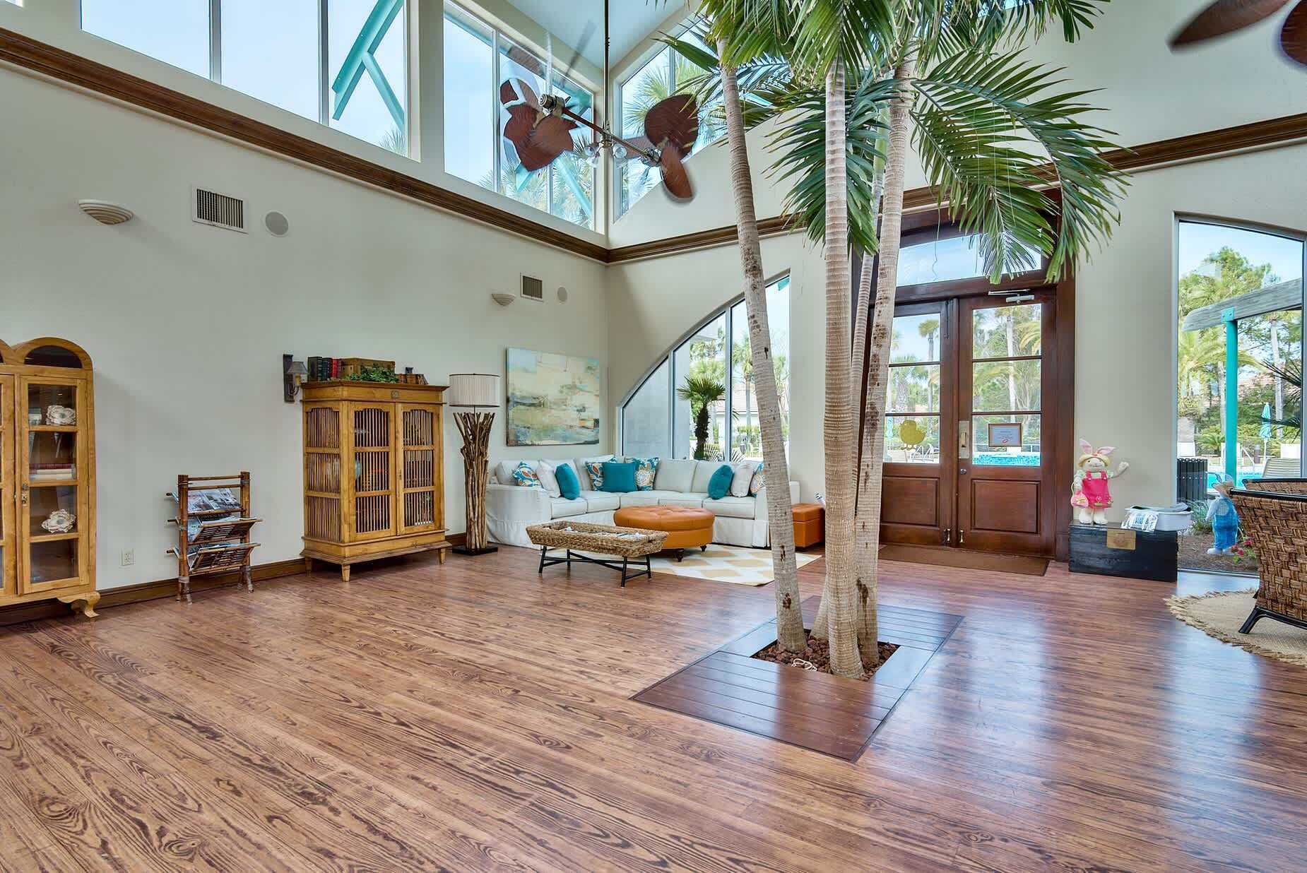 A spacious living room with a high ceiling, wooden flooring, a central seating area with a sofa and ottoman, a tall palm tree, and a large window providing natural light.