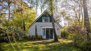 A small, single-story house with a green roof and white walls, surrounded by trees and a grassy yard.