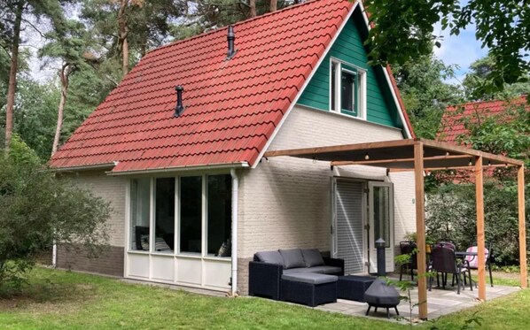 Et hus med rødt tegltag, en overdækket terrasse med udendørs møbler og et stort vinduesparti foran.