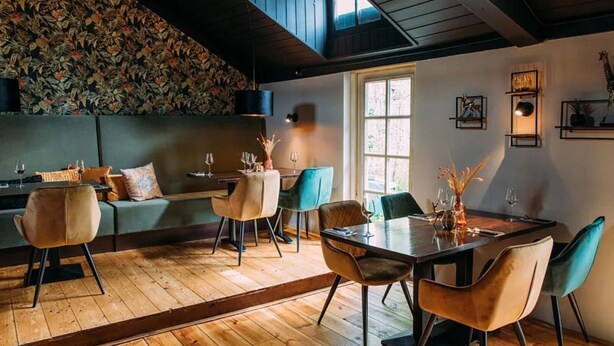 A dining area with a wooden floor, a high ceiling, and a large window. There are chairs and a table with a vase of flowers.