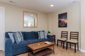 Un salon avec un canapé bleu, deux fauteuils en bois, une table basse en osier, un miroir et une affiche accrochée au mur.