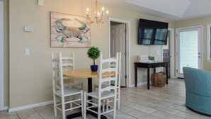 A dining area with a wooden table and white chairs, a chandelier, a wall-mounted TV, and a door leading to another room.