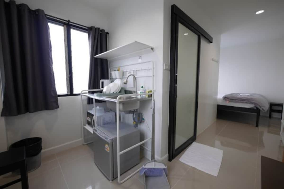 A hotel room with a bed, a small desk with a chair, a window with curtains, a bathroom with a shower, and a trash bin.