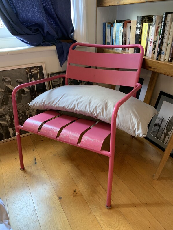 Una silla metálica rosa con cojín blanco en una habitación con suelo de madera, una estantería llena de libros y una ventana con cortinas.