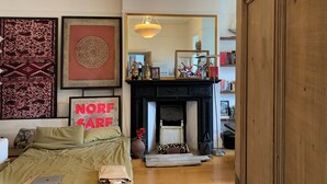 A cozy living room with a fireplace, a sofa, and a bed. There is a framed picture on the wall and a mirror reflecting the room.
