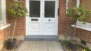 A white double door with glass panels, flanked by two potted plants, set against a brick wall with a metal decorative element above.