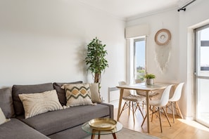 Ein Wohnzimmer mit einer Couch, einem Glastisch, einem Esstisch mit Stühlen, einer Pflanze und einem Wandbehang als Traumfänger.