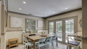 A dining area with a wooden table, light blue chairs, and a view through large glass doors. - Sunset Rentals | 5 Rice Lane | Dog Friendly (Hilton Head Island)