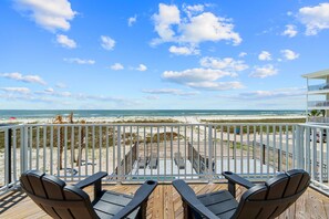 Une terrasse en bois avec deux chaises et une table, donnant sur une plage sous un ciel dégagé et parsemé de nuages.