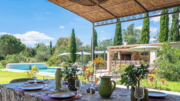 Un área de comedor al aire libre con una mesa dispuesta para cuatro personas, rodeada de abundante vegetación y una piscina al fondo.