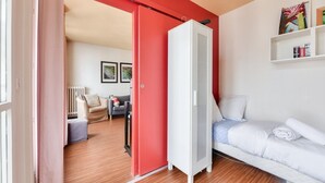 A room with a red door, a white bed, a grey sofa, and a white radiator. There are two framed pictures on the wall and a shelf with books. - Superb T3 near Place des Vosges (Paris)