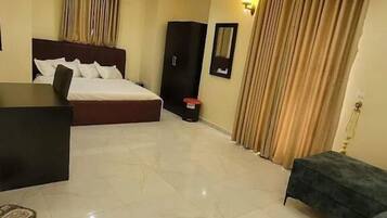A spacious hotel room with a large bed, a desk, and a sofa. The room has a tiled floor and a ceiling with recessed lighting.